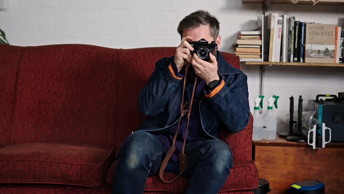 Photographer holding vintage camera to face while sitting on red couch in home studio.