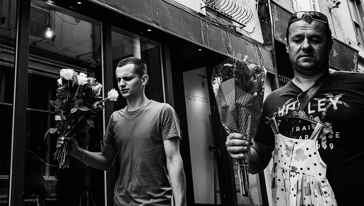 Two men on an urban street holding flower bouquets, photographed in black and white.