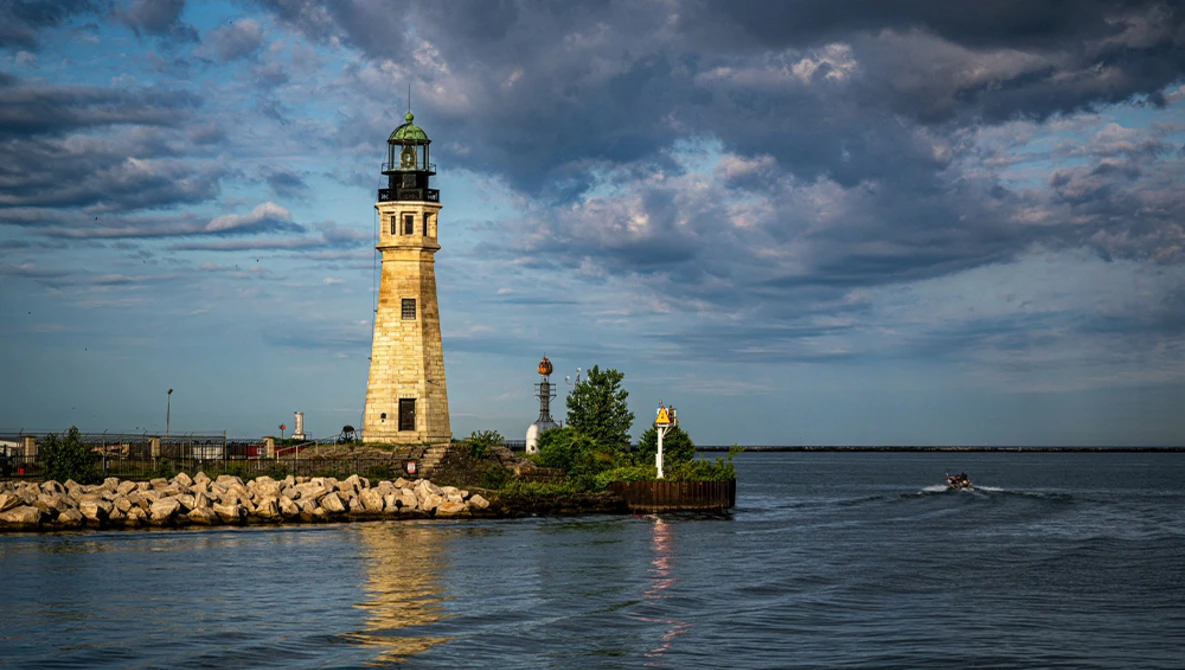 Stone lighthouse with green lantern room on rocky pier at sunset, dramatic cloudy sky reflected in calm water.