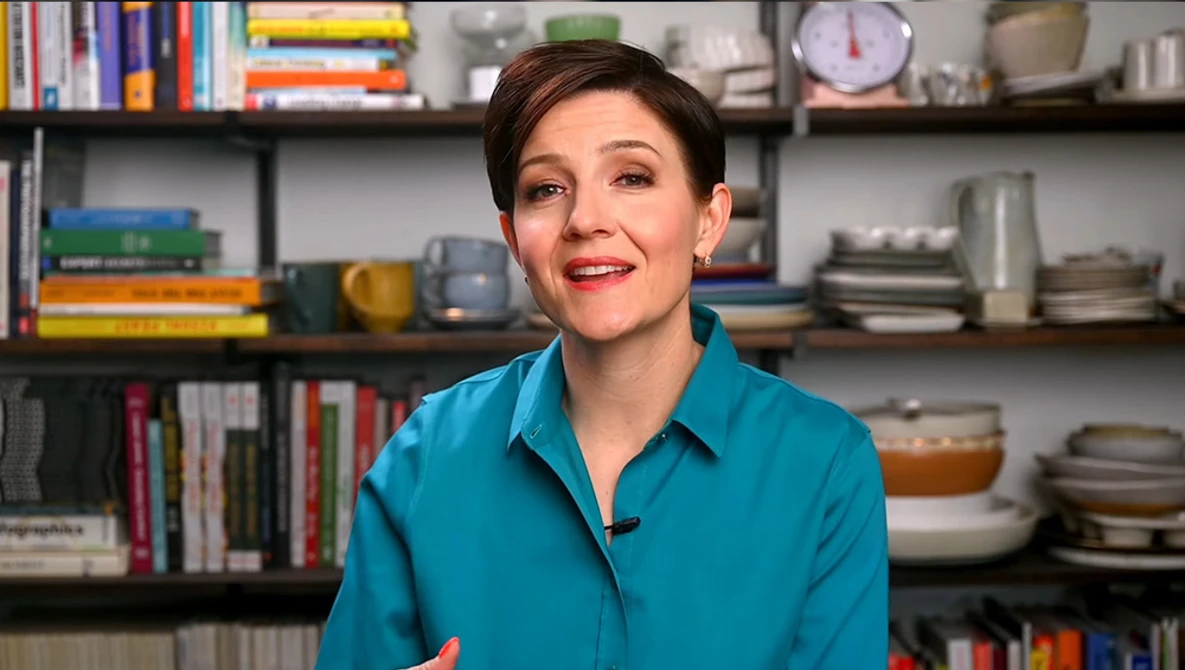 Woman in turquoise shirt speaking in front of shelving with books and kitchenware.