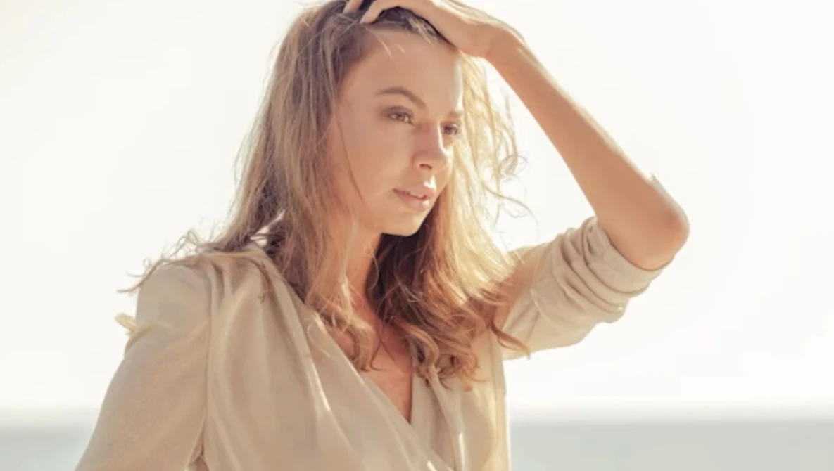 Woman in beige shirt with hand raised to head, backlit by golden sunlight against neutral background.