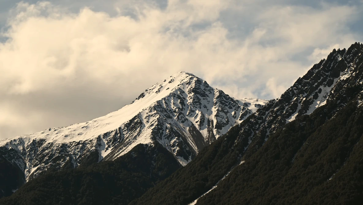 Snow-capped mountain peak with forested slopes under partly cloudy sky.