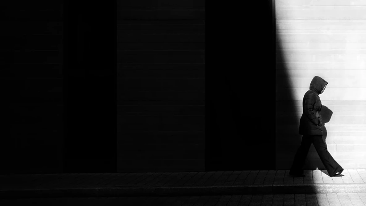 Silhouetted figure in dark clothing walking across a stark white floor against a black wall.