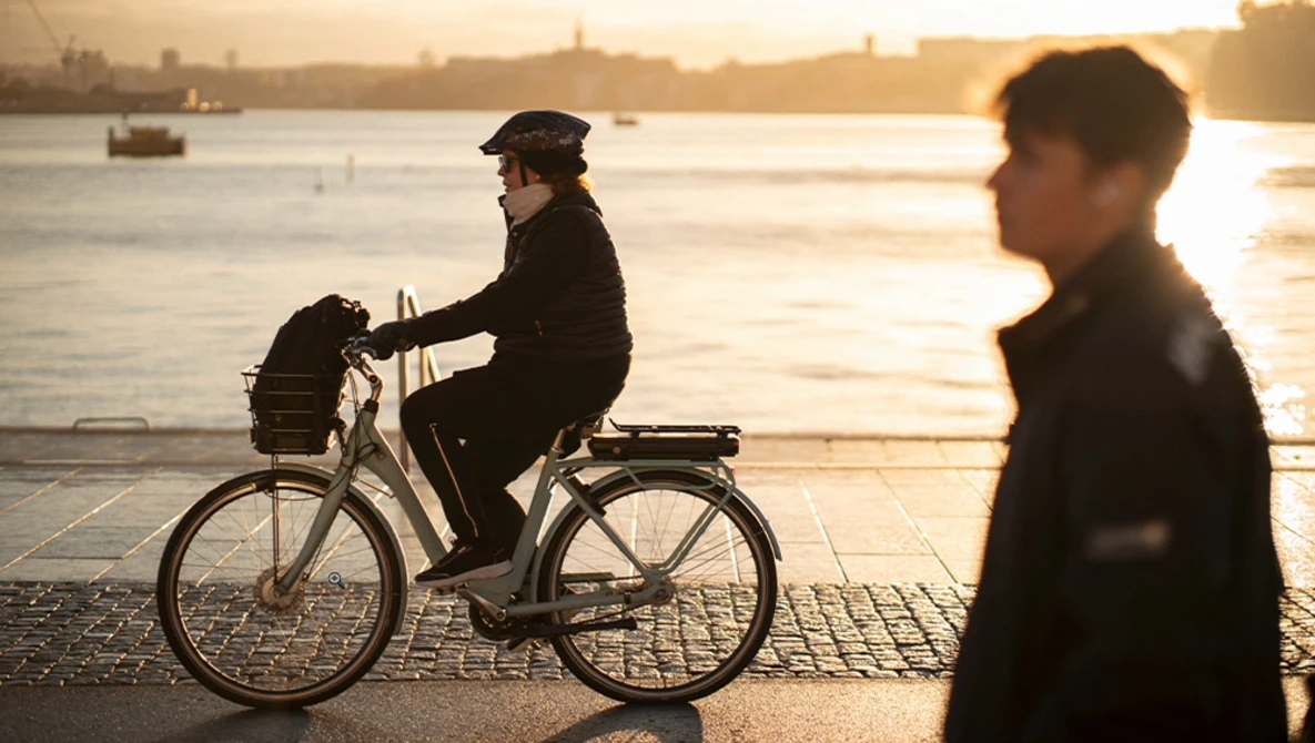 Person riding a bicycle on a waterfront promenade at golden hour with a blurred figure walking nearby.