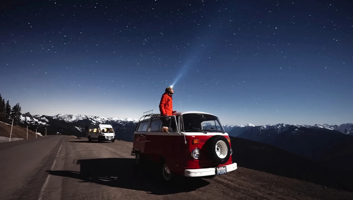 Person standing on roof of red and white vintage van at night, shining flashlight skyward with snow-capped mountains and starry sky in background.