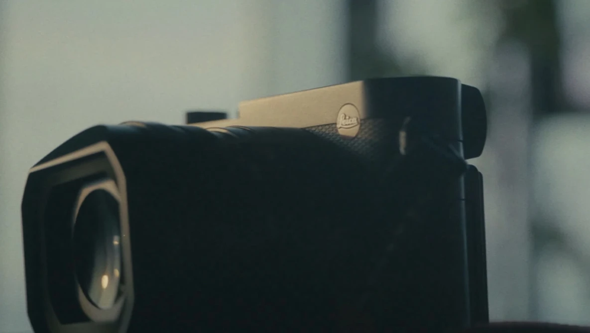 Black compact camera with a beige/tan hot shoe and visible lens, photographed against a soft blurred background.