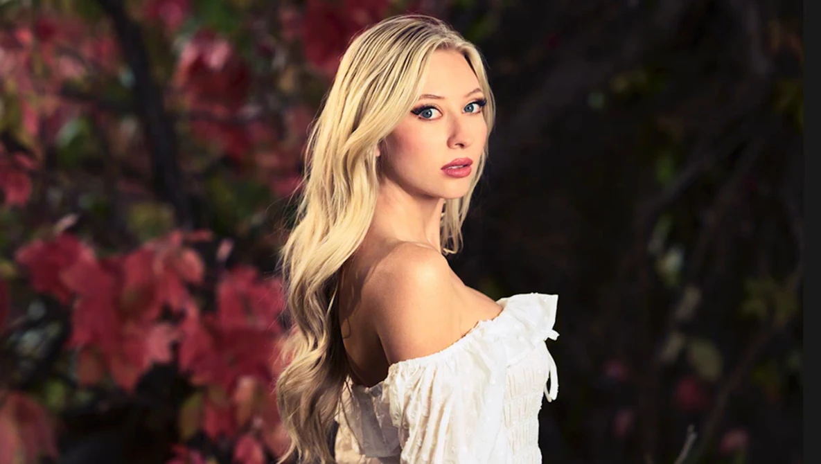 Woman with blonde wavy hair wearing an off-shoulder white ruffled top, photographed against a blurred background of pink flowering branches.