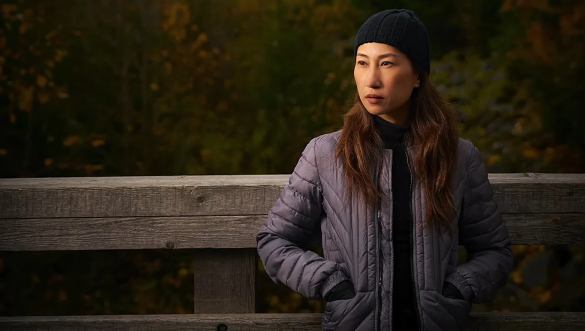 Woman in puffy jacket and black beanie sitting against wooden railing with blurred forest background.