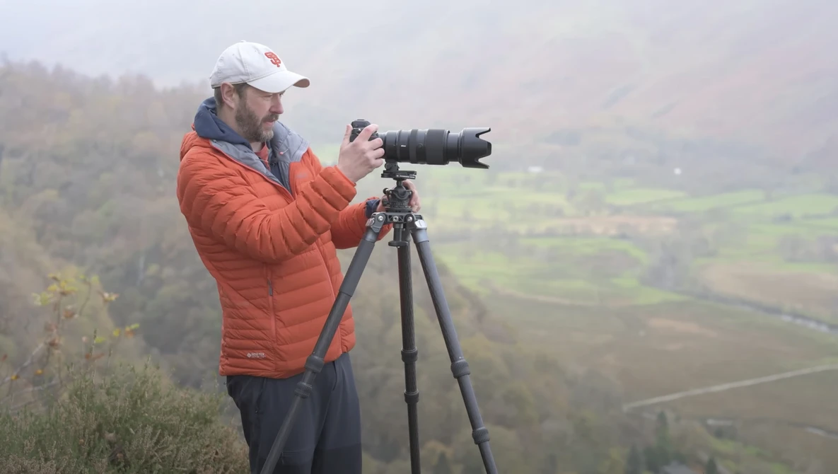 Photographer with telephoto lens mounted on tripod, positioned on elevated terrain overlooking misty valley landscape.