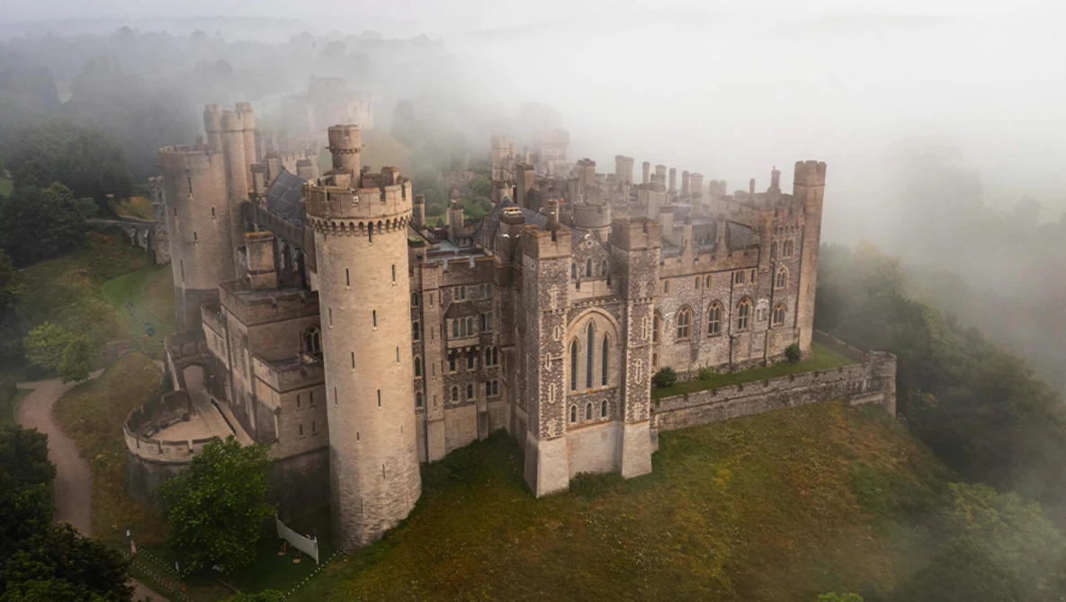 Medieval castle with multiple towers shrouded in mist, surrounded by forested hillside.