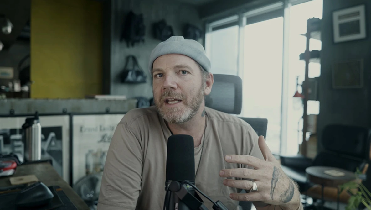 Man speaking into microphone in a home studio setup with casual attire and cap.