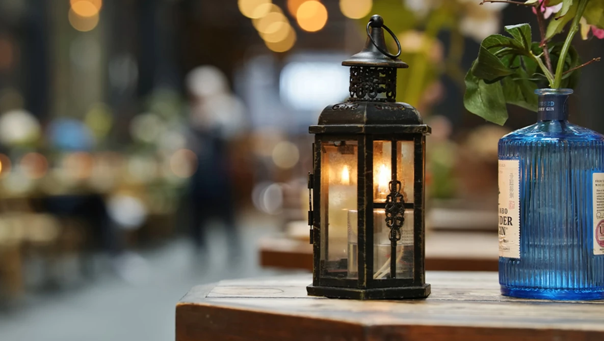 Black metal lantern with lit candle on wooden surface, flanked by blue glass bottle, with blurred bokeh lights in background.