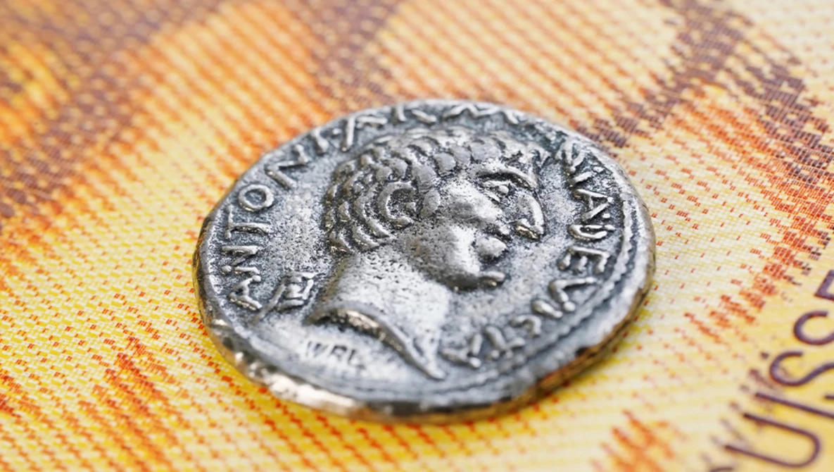 Ancient Roman coin with profile portrait resting on colorful euro banknotes.