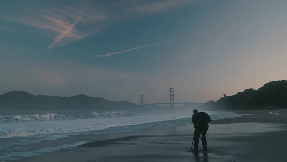 Person standing on wet beach at dusk with suspension bridge visible across the bay.