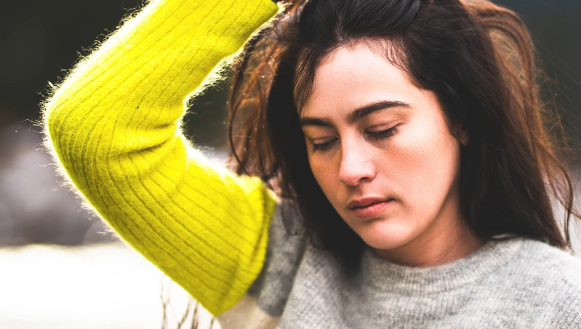 Woman with long dark hair wearing a bright yellow-green cardigan over a gray sweater, looking downward with a contemplative expression.