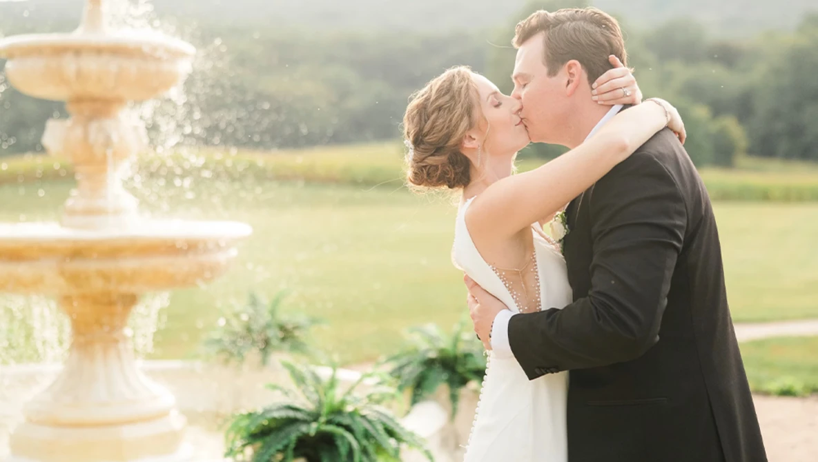 Bride and groom kissing by a fountain in a manicured garden setting.
