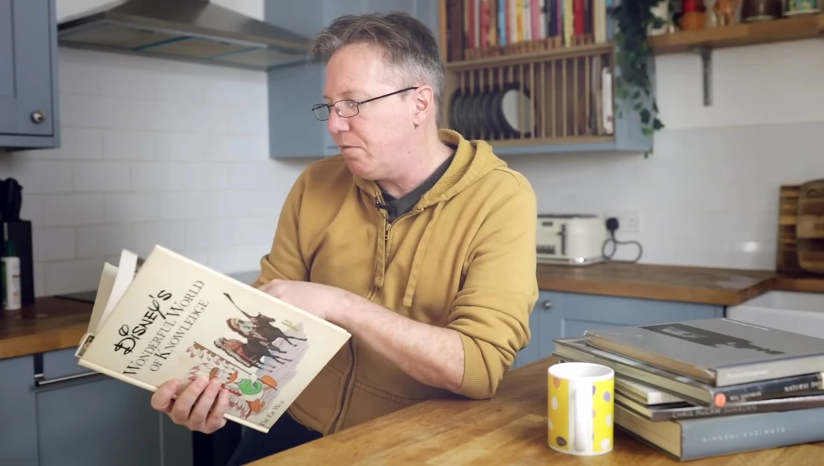 Man in yellow cardigan examining a printed publication at a wooden desk in a kitchen.