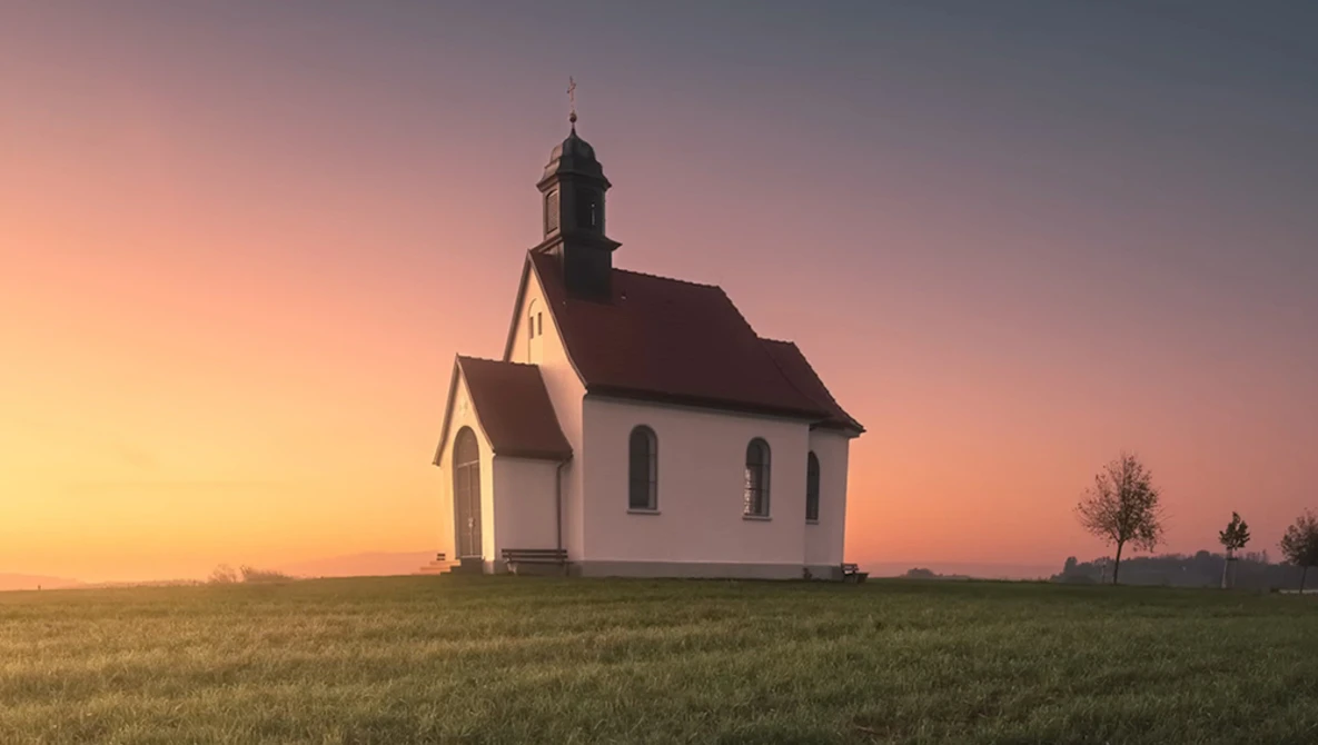 White wooden church with dark roof standing alone in a vast prairie at sunset.