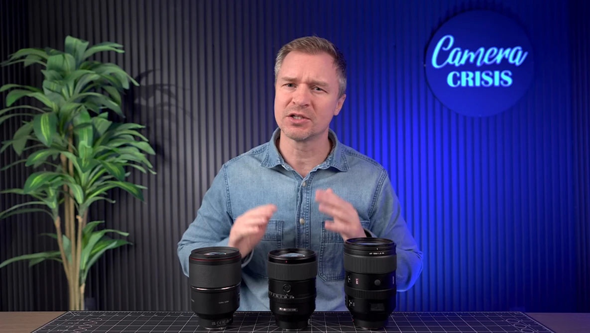 Man in denim shirt demonstrating three camera lenses on a table in a studio setting.