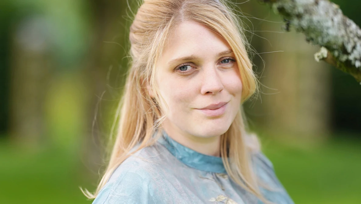 Young woman with blonde hair looking at camera in outdoor setting with green background.