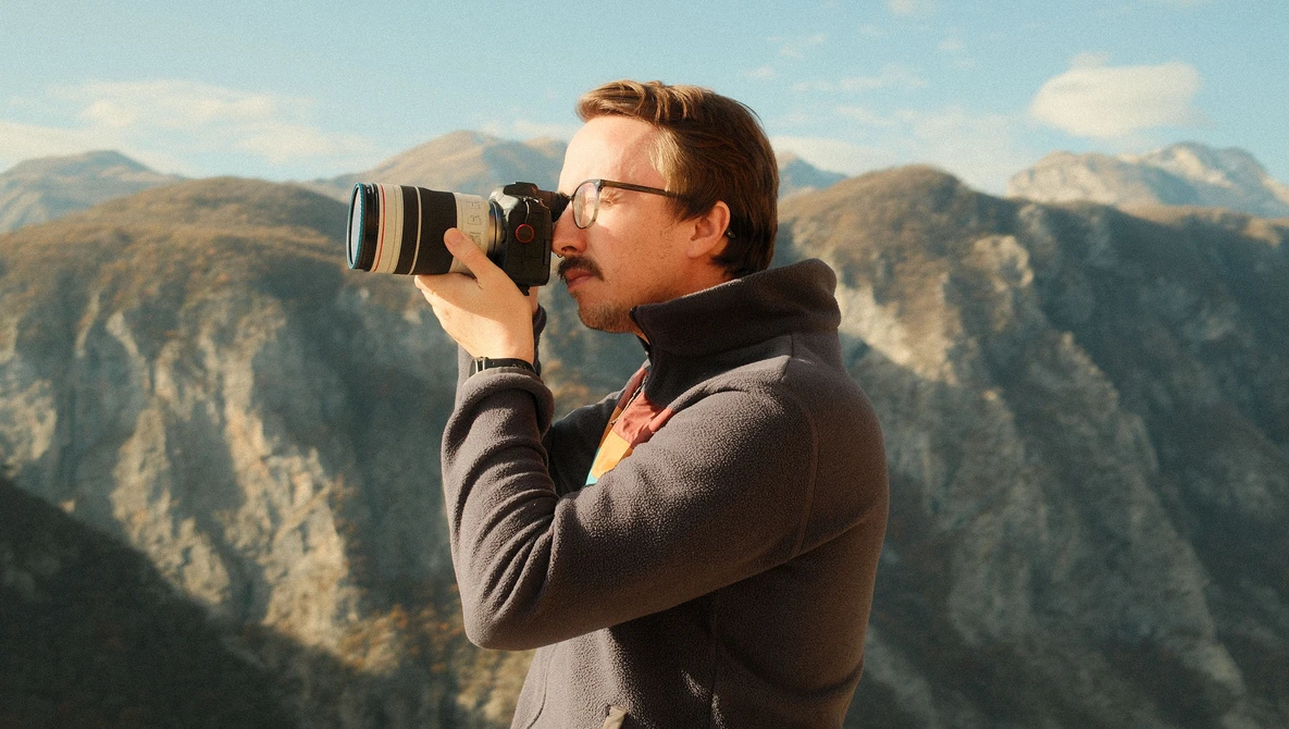 Photographer with glasses holding a DSLR camera with telephoto lens, shooting in a mountainous landscape.
