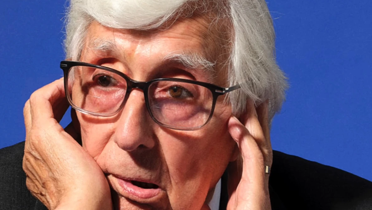 Older man with white hair and glasses against a blue background, hand near his face in a contemplative pose.
