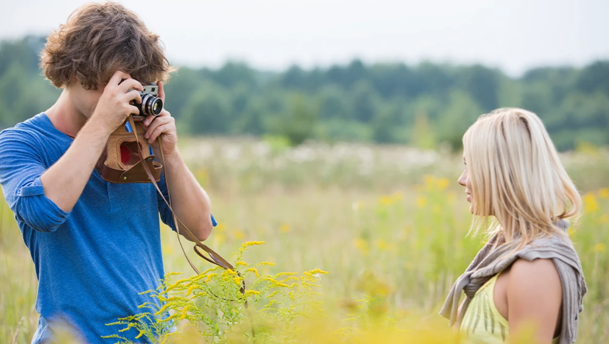 Young man with curly brown hair photographing a blonde woman in a sunlit field with vintage camera.
