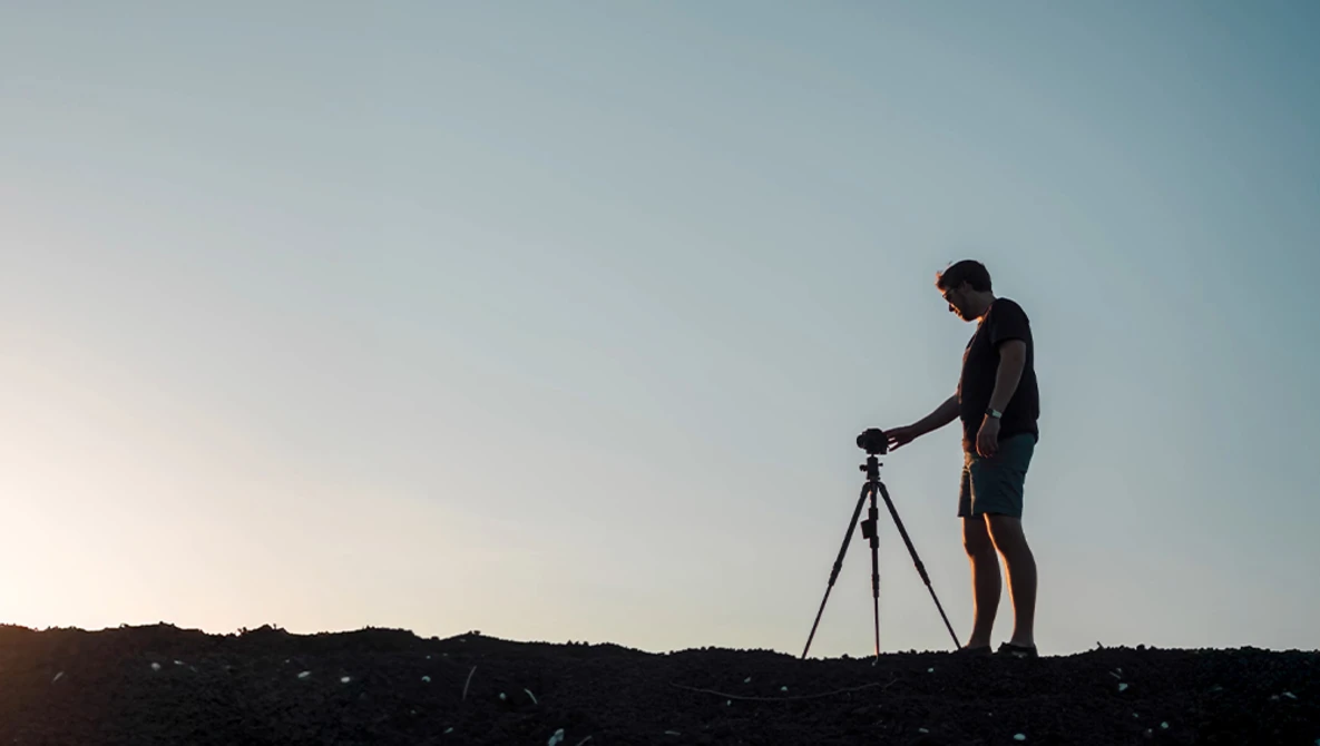 Silhouetted photographer operating a tripod-mounted camera against a clear sky at dusk.