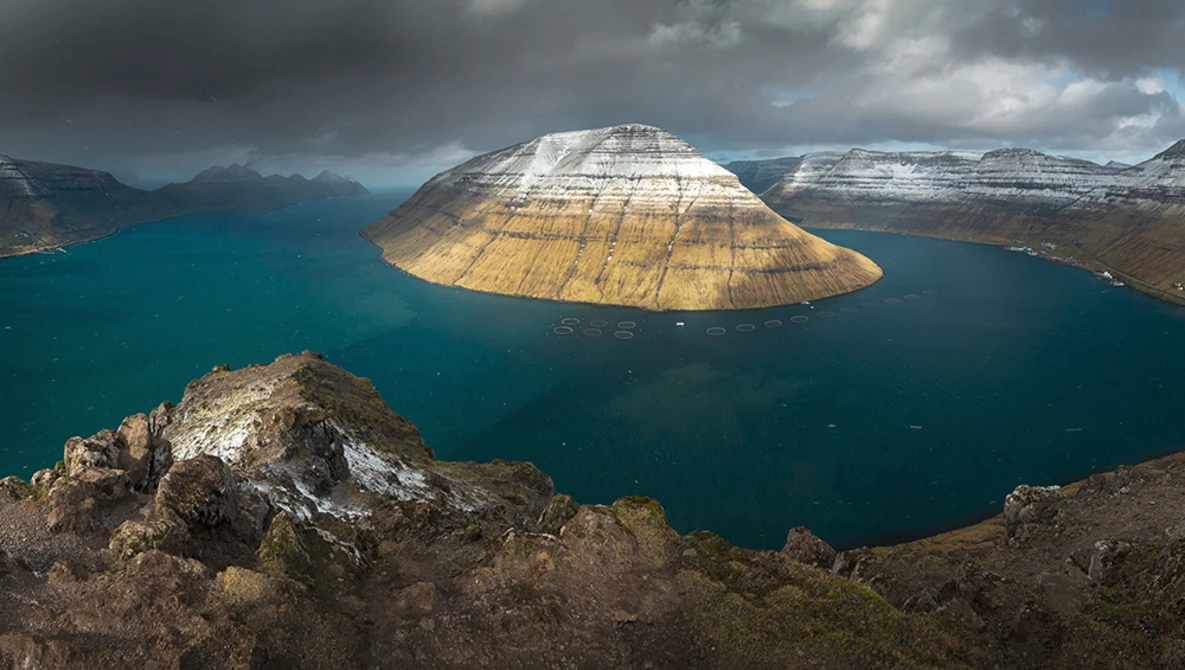 Snow-capped mountain with distinctive dome shape rising from turquoise fjord waters, viewed from rocky foreground.