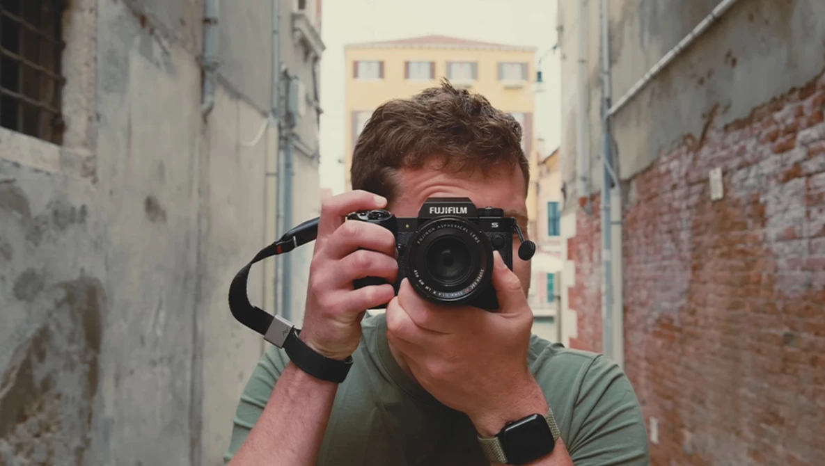 Photographer holding a Fujifilm mirrorless camera up to his face in a narrow urban alleyway.
