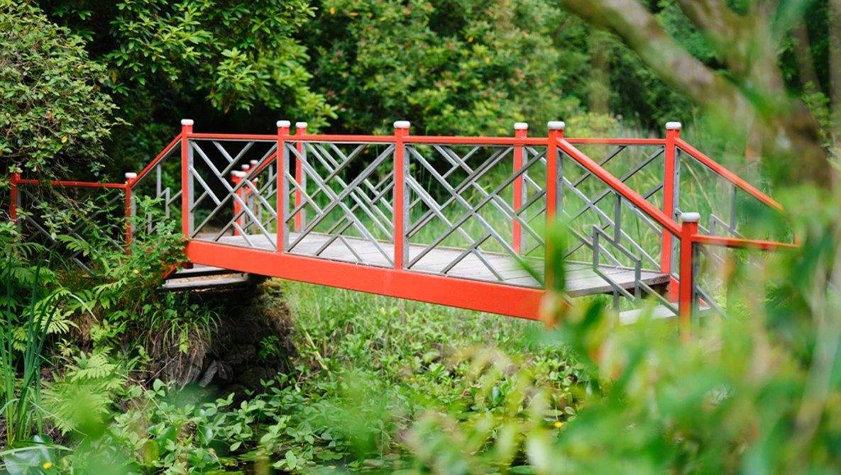 Bright red footbridge with X-pattern railings spanning a small stream in a lush garden.