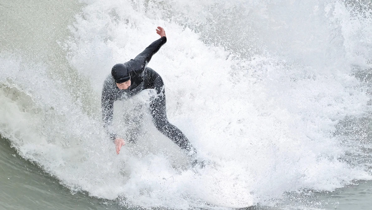 Surfer in black wetsuit riding a large wave with significant white water and spray.