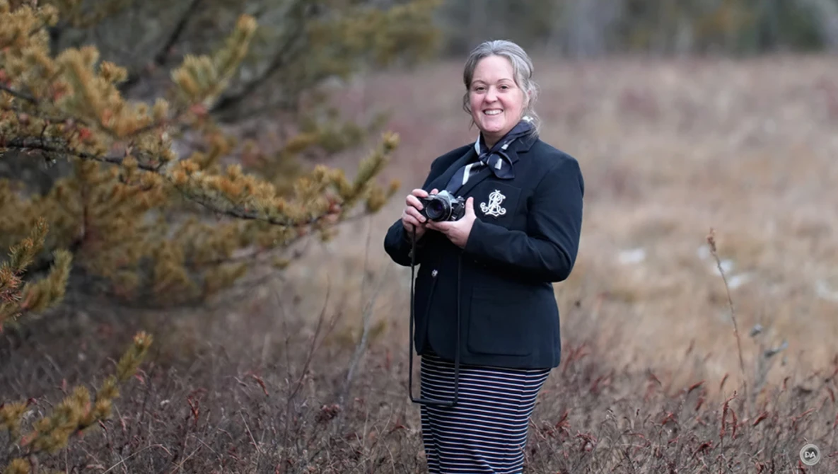 Female photographer holding a camera in an open field with sparse vegetation and evergreen shrubs.