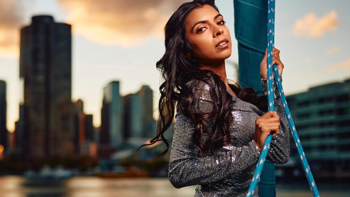 Woman in sequined top holding blue metal beam against urban skyline at sunset.