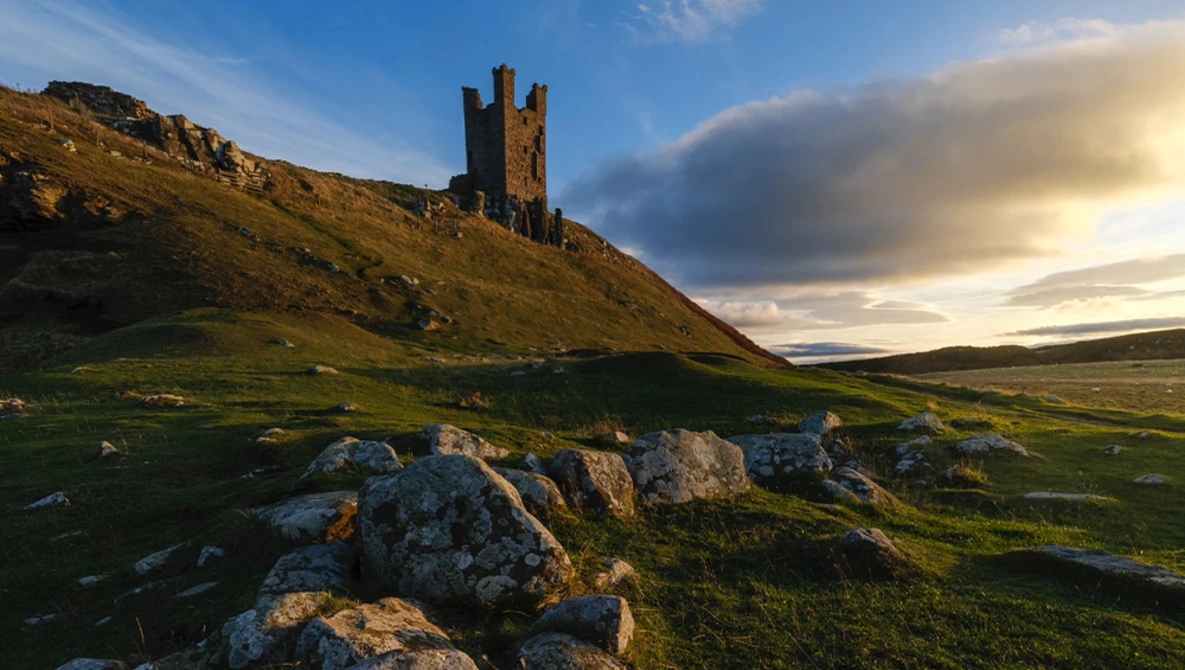 Medieval stone tower ruins on a hillside with golden hour sunlight and dramatic clouds.