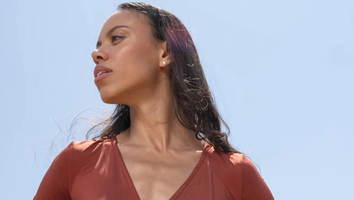 Woman with long dark hair wearing rust-colored top, photographed in profile against clear blue sky.
