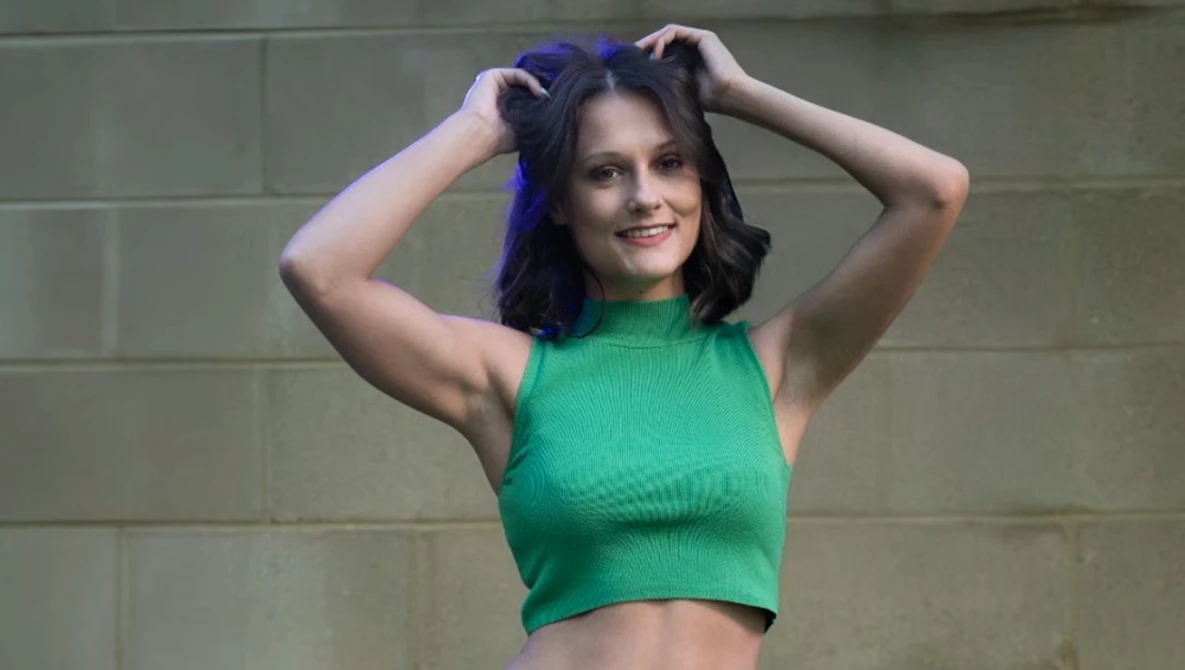Woman in green crop top with dark blue hair against neutral brick wall background.