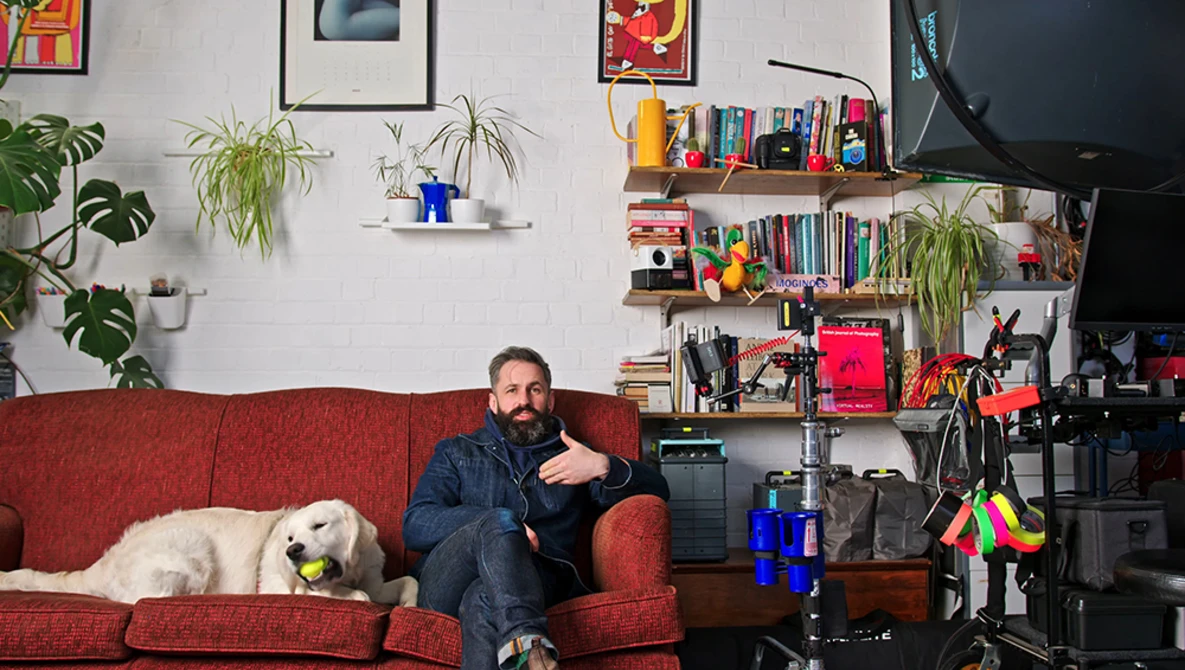 Man sitting on red couch with white dog in home studio setup with shelving and camera equipment.