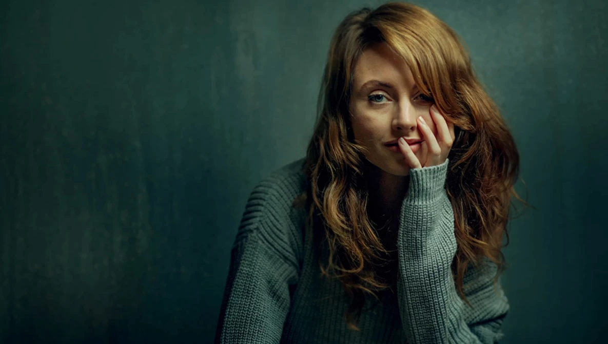 Woman with reddish-brown wavy hair wearing a sage green cardigan, resting her face against her hand against a muted teal background.