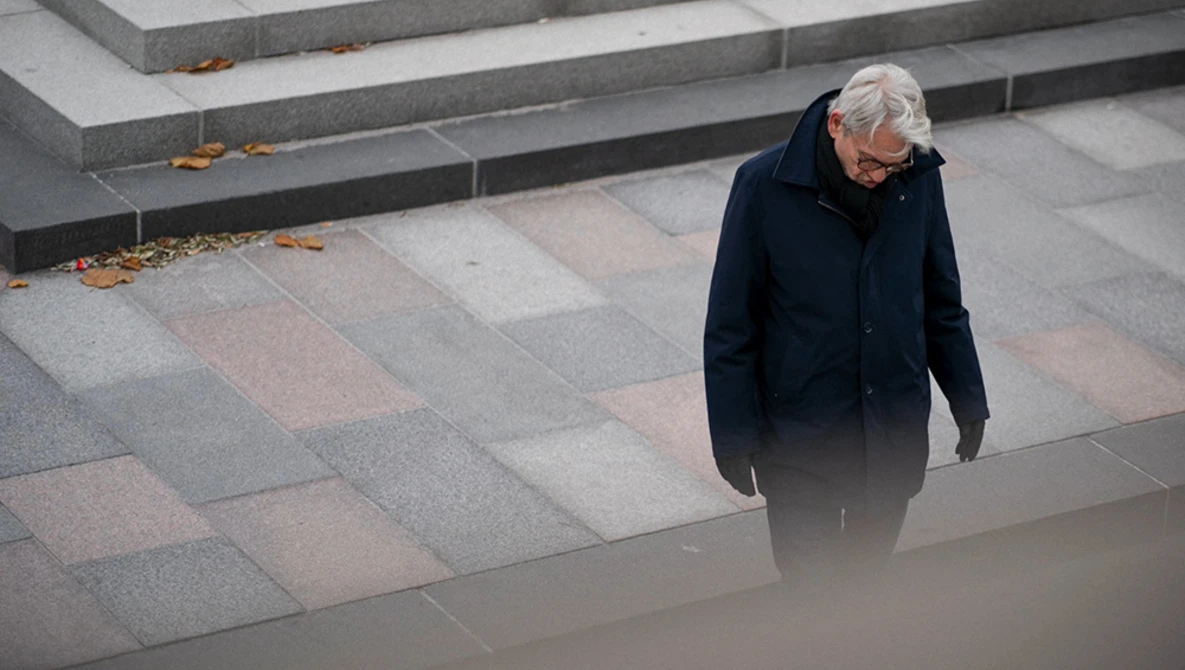 Elderly person in dark coat walking alone across geometric paved plaza with stepped levels.