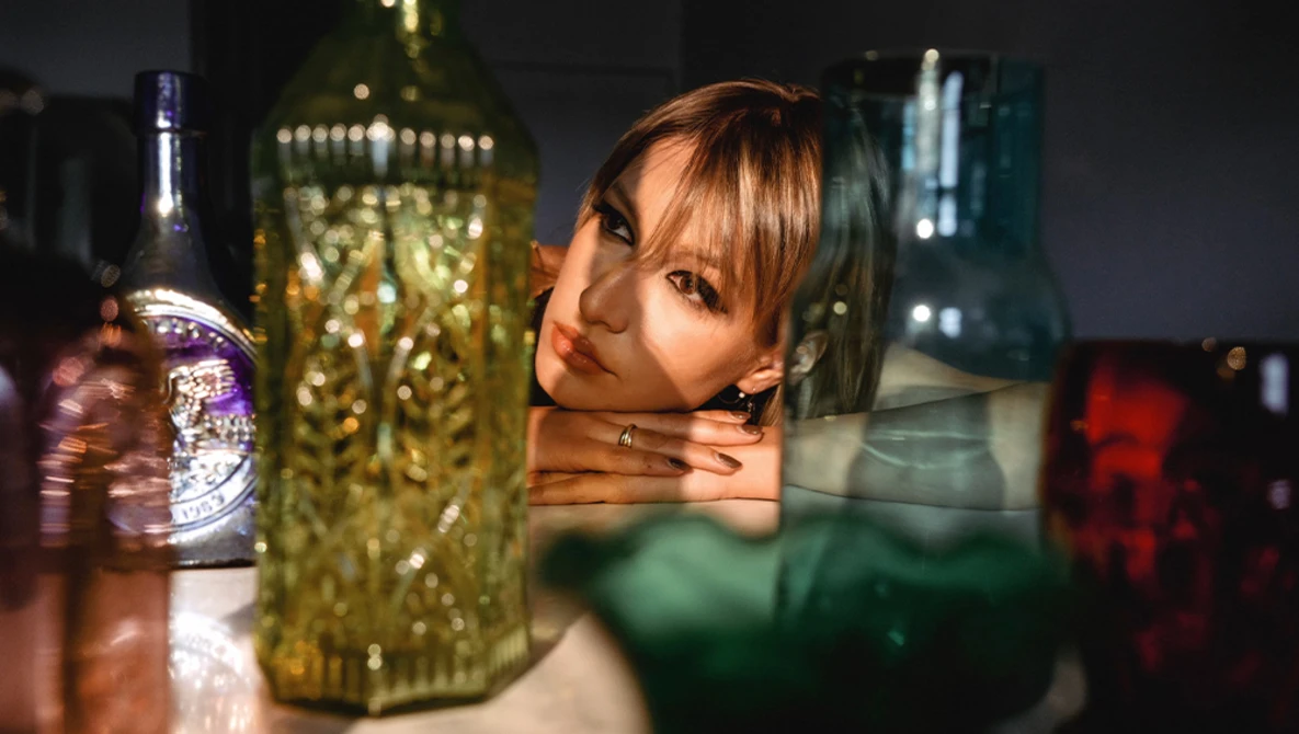 Woman's face reflected in glass bottles with warm studio lighting.