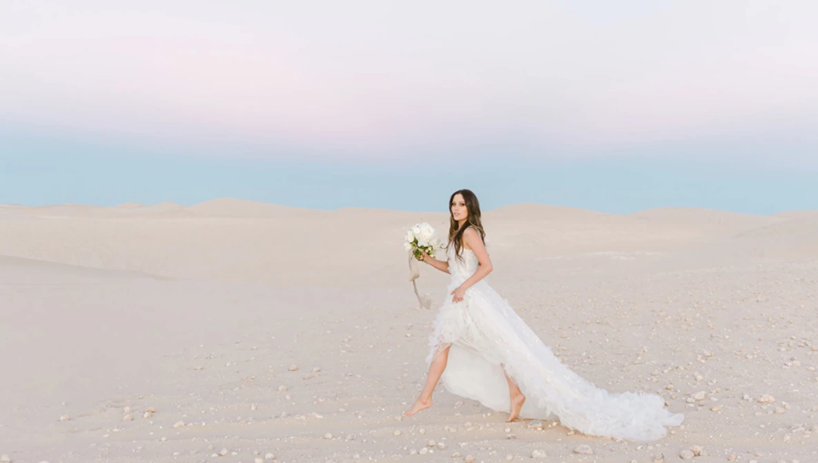 Bride in flowing white gown holding bouquet on sandy beach at dusk.