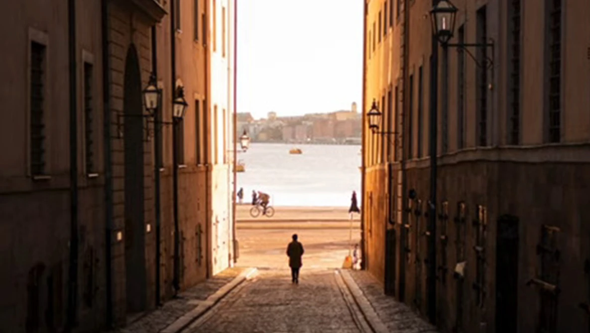 Narrow cobblestone alley between terracotta buildings framing a waterfront view with a solitary figure walking toward the water.