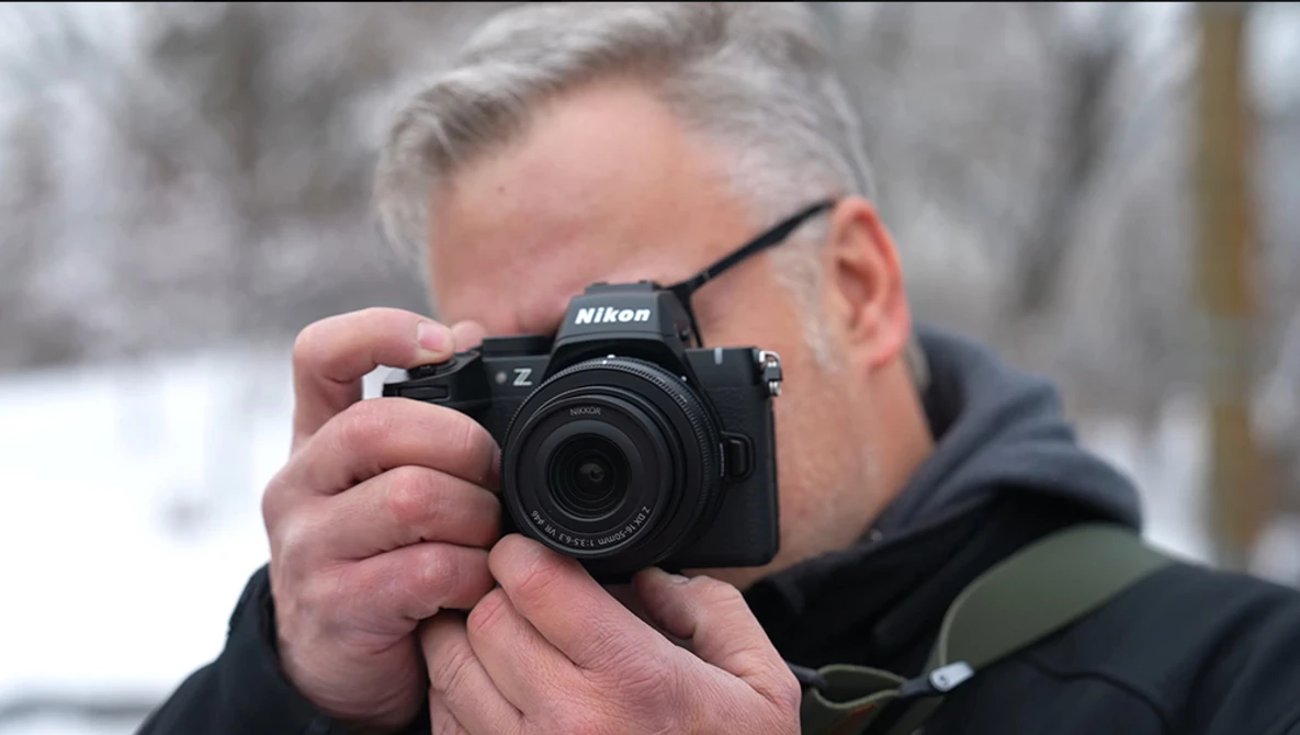 Photographer holding a Nikon Z mirrorless camera up to his face while wearing glasses and dark winter clothing.