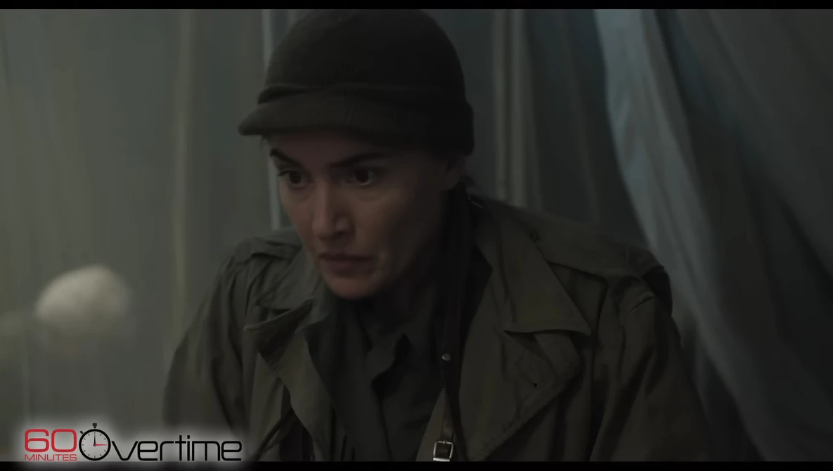 Person wearing dark fedora hat and olive jacket in dimly lit interior space.