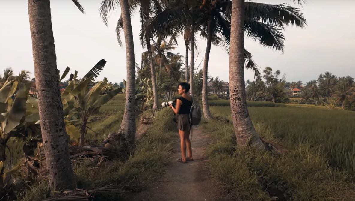 Woman walking on a dirt path between palm trees with rice fields visible in the background.