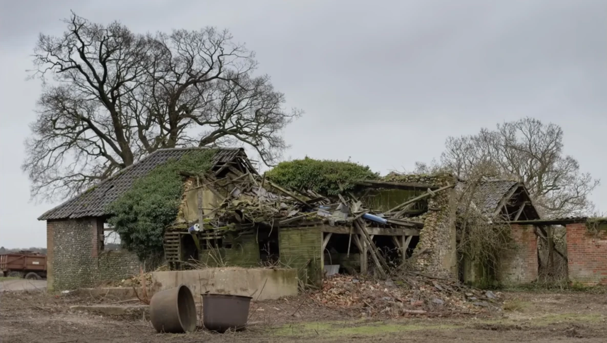 Abandoned stone cottage overgrown with moss and vegetation, surrounded by bare trees and a metal drum in the foreground.