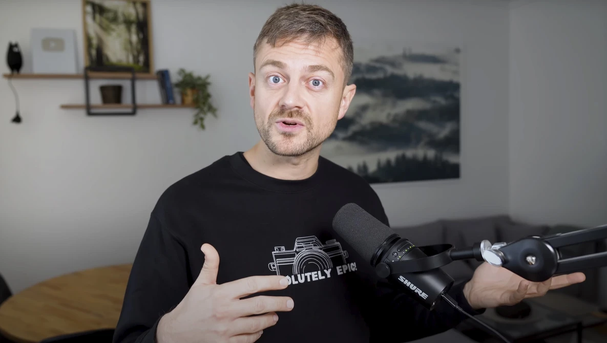 Man in black camera-themed t-shirt speaking expressively while holding a microphone in an indoor studio setting.
