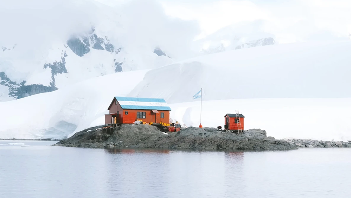 Bright orange research station building on a rocky outcrop in polar waters, with snow-covered mountains and glaciers in the background.