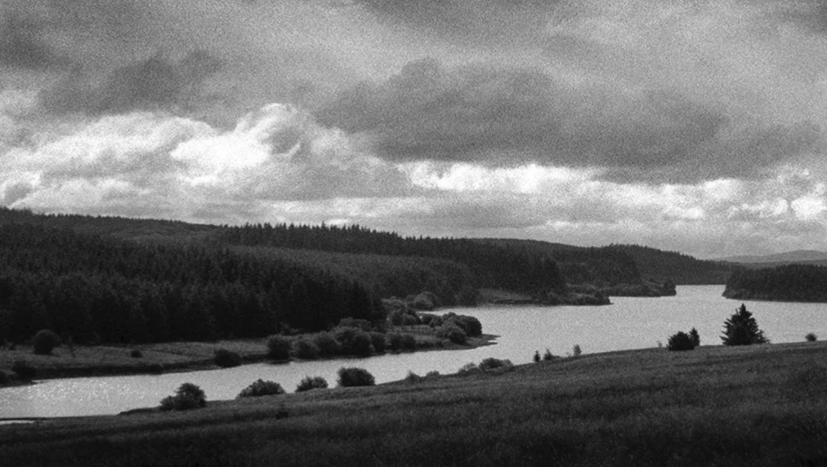 Dramatic landscape with forested hills, a calm lake, and storm clouds overhead in black and white.